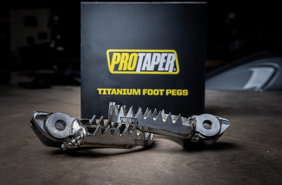 ProTaper Titanium Foot Pegs displayed in front of branded packaging, highlighting their durable construction, aggressive teeth for enhanced grip, and sleek titanium finish.